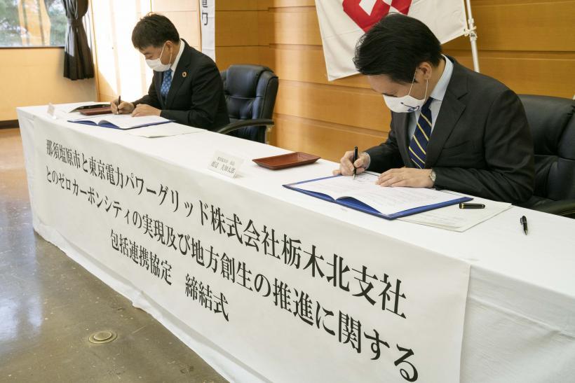二人の男性が、締結式の垂れ幕がかかった長机の上で書類にサインをしている写真