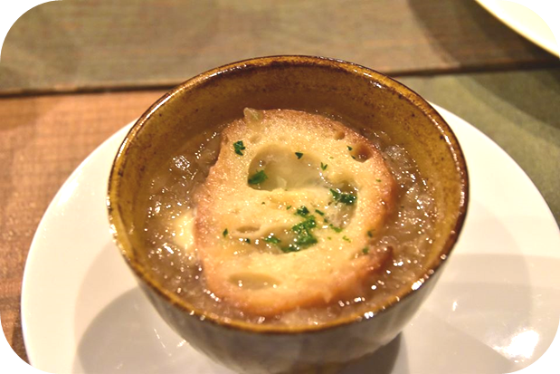 白いお皿に乗せられた茶色い器にパンが浮かべられたオニオングラタンスープの写真