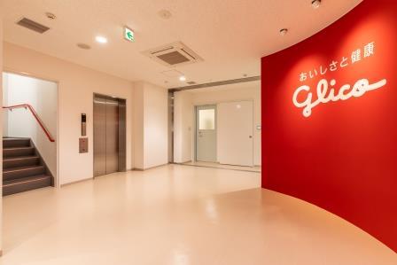 右においしさと健康 glicoと書かれた赤い壁があり、階段やエレベーターがある室内の写真