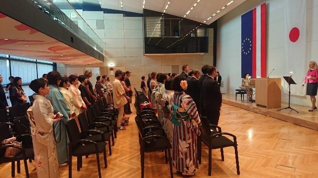 日本の国旗とオーストリアの国旗が並んだホールで参加者が起立している様子の写真