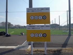 青木サッカー場Cグラウンド外縁に立つ標識2枚の写真。標識にはそれぞれスポーツ振興くじ助成金を受けて整備された旨が書かれているのが確認できる