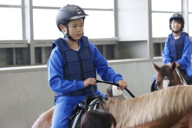 乗馬体験にて騎乗する児童の写真。体験に臨んで乗馬用ヘルメットとプロテクターを各自着用しているのが確認できる