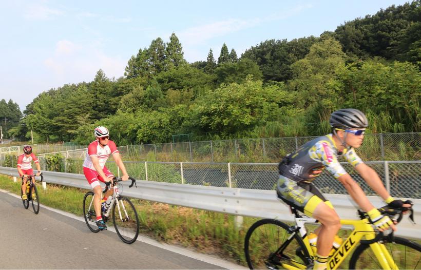小川沿いの車道脇を一列になってロードバイクで走るオーストリア選手たちの写真