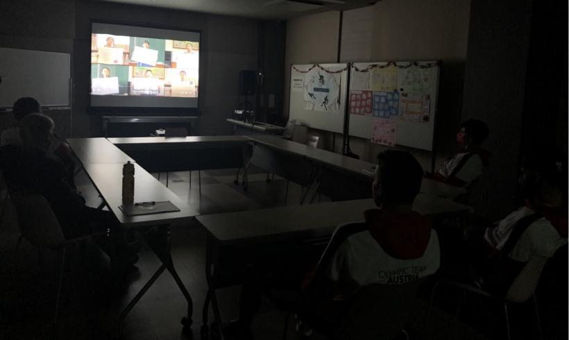 照明を落とした会議室でスクリーンに映し出された動画を鑑賞するオーストリア選手たちの写真。スクリーンにはメッセージボードを見せつつ合唱する児童たちのカットが映し出されているのが確認できる